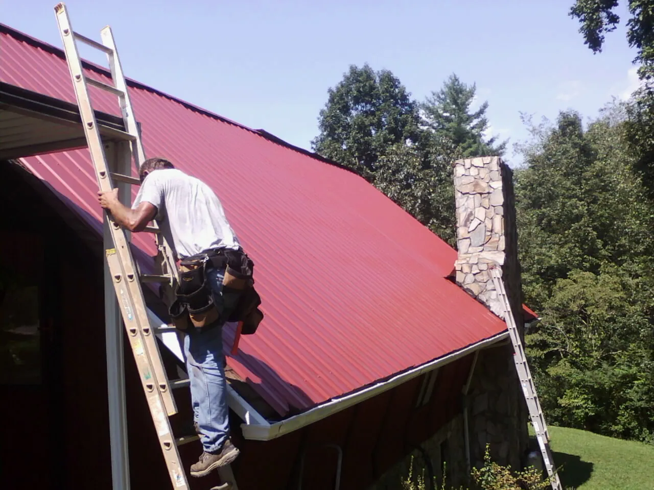 Roofer on a ladder beside a red metal roof and stone chimney — Soffit & Fascia Repair by Roofing Brothers in West Haven